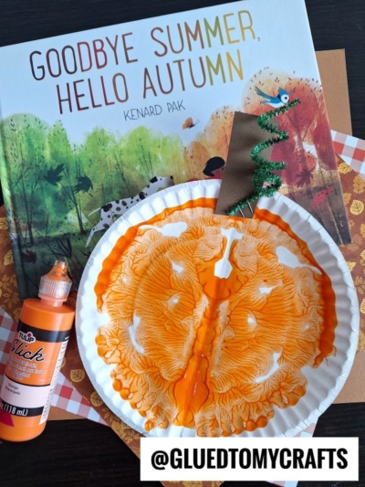 A children’s book titled “Goodbye Summer, Hello Autumn” lies under a fall-themed paper and a paint splat pumpkin paper plate craft with orange paint, a brown paper stem, and a green pipe cleaner vine. An orange glue bottle is beside them.