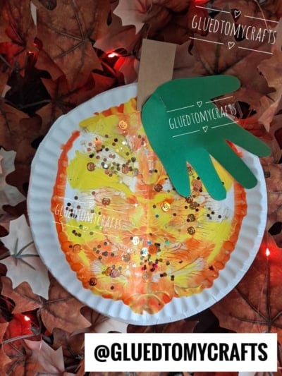 A paint splat pumpkin craft made from a paper plate with orange and yellow paint, glitter, a green handprint leaf, and a brown stem, surrounded by artificial autumn leaves.