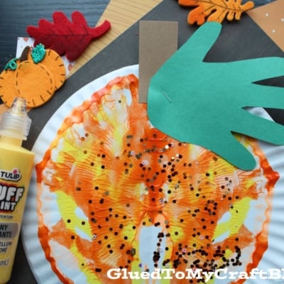 A paper plate with orange and yellow paint splatters resembles a paint splat pumpkin, decorated with black glitter, a green paper stem, and a green cutout “leaf.” Autumn-themed felt decorations surround it.