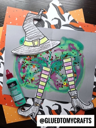 A Halloween craft featuring a witch’s hat and legs made from colored paper, glitter, and paint splat details for a fun melted witch effect, with a bottle of turquoise paint nearby and festive background papers. The handle @GLUEDTOMYCRAFTS is displayed.