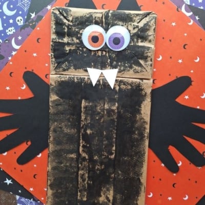 A paper bag decorated as a black cat with googly eyes, paper fangs, and black handprint cutout wings sits on colorful Halloween-themed paper. Perfect for Halloween Crafts for Kids! A label reads @GLUEDTOMYCRAFTS.