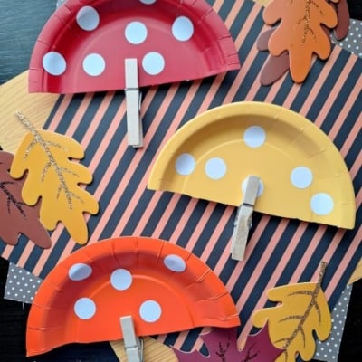 Three paper plate mushrooms made from red, orange, and yellow plates with white spots and clothespin stems, placed on autumn leaf and striped paper decorations—perfect FALL CRAFTS FOR KIDS! @GLUEDTOMYCRAFTS is at the bottom.