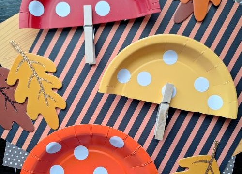 Three paper plate mushrooms made from red, orange, and yellow plates with white spots and clothespin stems, placed on autumn leaf and striped paper decorations—perfect FALL CRAFTS FOR KIDS! @GLUEDTOMYCRAFTS is at the bottom.