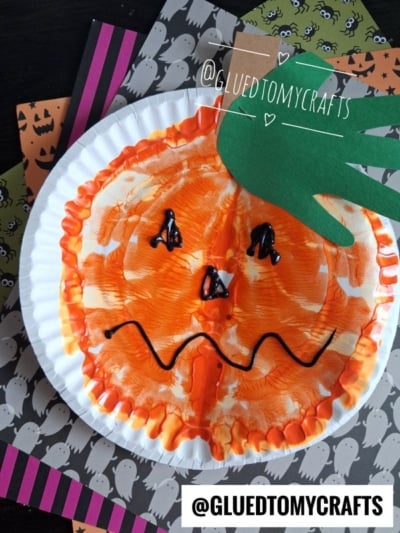 A paint splat pumpkin paper plate craft with orange paint and a green paper hand stem, placed on Halloween-themed paper. The pumpkin has a black painted face and the handle @GLUEDTOMYCRAFTS is displayed.