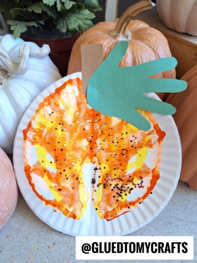 A paper plate painted orange and yellow in a playful paint splat pumpkin style, decorated with green and brown paper for a stem and leaf. The plate is surrounded by real pumpkins. Text at the bottom reads @GLUEDTOMYCRAFTS.