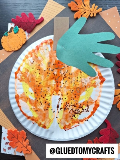 A paint splat pumpkin made from a paper plate, decorated with orange and yellow paint to resemble a pumpkin, is topped with a green paper handprint stem and surrounded by felt leaves and other pumpkin crafts on a dark surface.