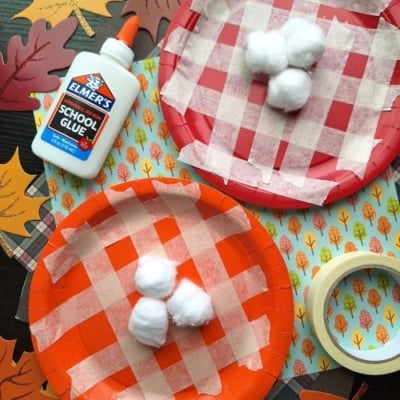 Two paper plates with red and orange plaid patterns made from masking tape, each with three cotton balls glued on top—perfect for FALL CRAFTS FOR KIDS. Elmer’s glue, fall-themed paper leaves, and masking tape are also visible.