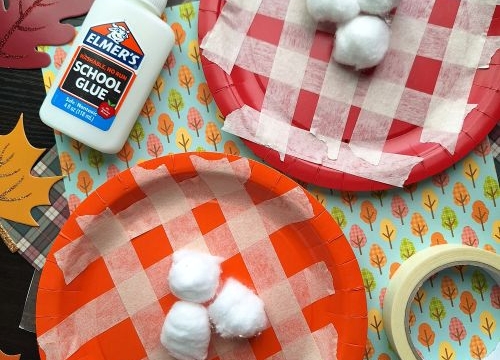 Two paper plates with red and orange plaid patterns made from masking tape, each with three cotton balls glued on top—perfect for FALL CRAFTS FOR KIDS. Elmer’s glue, fall-themed paper leaves, and masking tape are also visible.