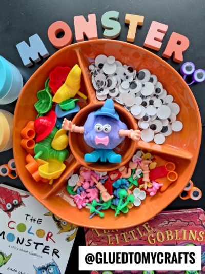 An orange tray holds colorful plastic monster parts, googly eyes, tweezers, and a clay monster figure—a creative addition to your FALL THEMED SENSORY BIN IDEAS. Surrounding the tray are books, plastic scissors, and large letters spelling “MONSTER.”.