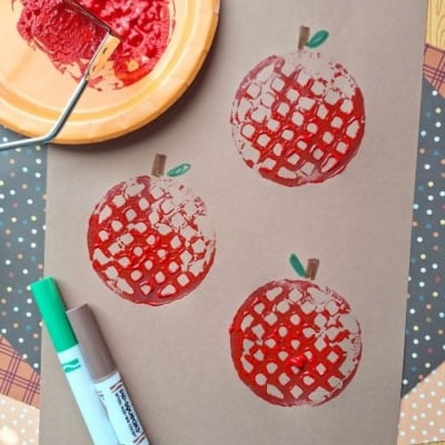 Brown paper showcases three red apple prints, created using a potato masher stamped craft technique. A green and brown marker and a plate with red paint sit nearby. The username @GLUEDTOMYCRAFTS is labeled at the bottom.