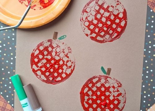 Brown paper showcases three red apple prints, created using a potato masher stamped craft technique. A green and brown marker and a plate with red paint sit nearby. The username @GLUEDTOMYCRAFTS is labeled at the bottom.