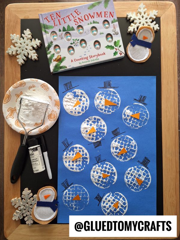 A children’s snowman craft set on a table: a “Ten Little Snowmen” book, blue paper with potato masher stamped craft snowmen, a plate with white paint and roller, felt snowman crafts, and snowflake decorations. @GLUEDTOMYCRAFTS is written at the bottom.