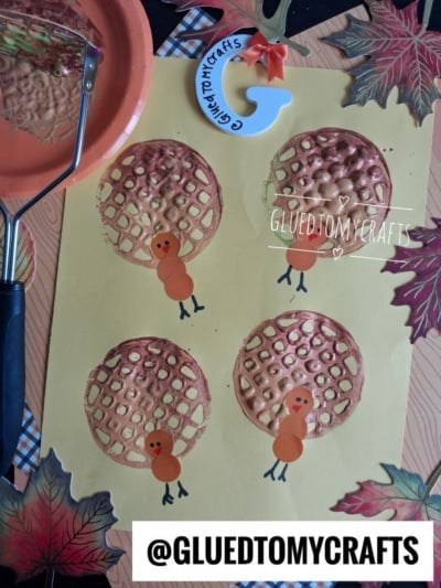 Four turkey crafts made from brown painted waffle prints, inspired by the classic potato masher stamped turkey technique, with added orange paper circles for heads and bodies, black marker legs, and eyes. Craft tools, fall leaves, and the @GLUEDTOMYCRAFTS logo surround them.