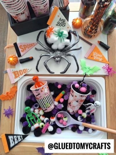 A festive Halloween craft setup with cups, napkins, and decorations featuring spiders, skeletons, bats, and colorful pom-poms—perfect inspiration for FALL THEMED SENSORY BIN IDEAS. Instagram handle @GLUEDTOMYCRAFTS is displayed at the bottom.