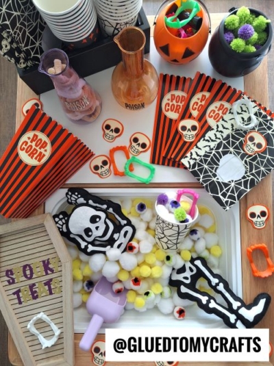 A Halloween-themed craft setup with striped popcorn bags, candy, mini cauldrons, skeleton decorations, plastic rings, fuzzy pom-poms—perfect inspiration for FALL THEMED SENSORY BIN IDEAS. The handle @GLUEDTOMYCRAFTS is displayed on a letterboard reading “SPOOKY TREATS.”.