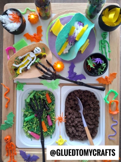 A Halloween-themed sensory play tray inspired by FALL THEMED SENSORY BIN IDEAS, featuring plastic skeletons, bats, insects, colorful shredded paper, and pretend food served with skeleton-hand tongs. Corner handle: @GLUEDTOMYCRAFTS.