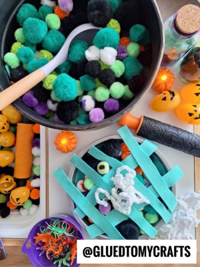 A variety of Halloween-themed craft supplies, perfect for fall themed sensory bin ideas, including colorful pom-poms in a pot, felt strips, pipe cleaners, toy spiders, small pumpkins, jack-o-lantern eggs, and a cauldron arranged on a table.