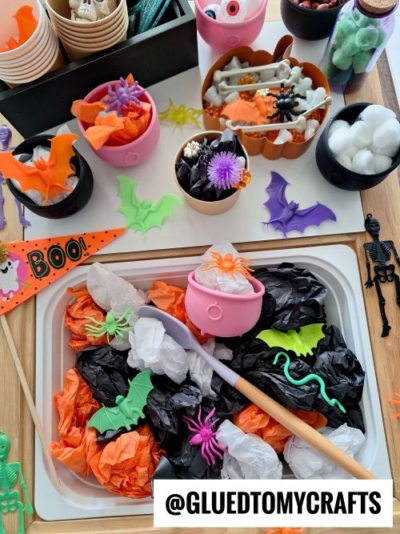 A Halloween-themed craft table with cauldrons, orange and black tissue paper, plastic spiders, bats, skeletons, and googly eyes—perfect for spooky crafting or as inspiration for FALL THEMED SENSORY BIN IDEAS. @GLUEDTOMYCRAFTS text at the bottom.