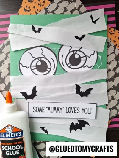 A torn paper mummy craft made with green paper, large cartoon eyes, white bandage strips, black bat cutouts, and a sign reading SOME MUMMY LOVES YOU! Elmer's glue and Halloween-patterned paper complete the spooky scene.