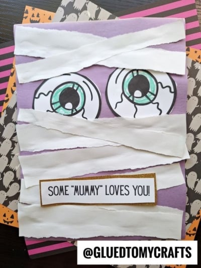 A Halloween card decorated as a torn paper mummy with googly eyes and layered strips, featuring a label that reads SOME MUMMY LOVES YOU! The background showcases striped and ghost-patterned paper for an extra festive touch.