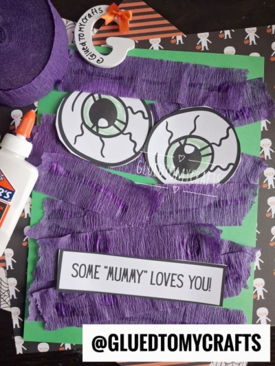 A Halloween craft features large cartoon eyes, purple crepe paper, Some Mummy Loves You! text, and a bottle of Elmers glue on a green background. This torn paper mummy design is perfect for spooky fun. @GLUEDTOMYCRAFTS is labeled at the bottom.