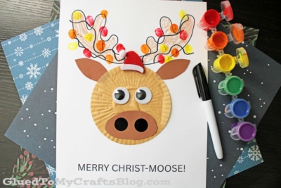 A cupcake liner moose head design, in the center of white cardstock paper, covered with Christmas lights made from a child's thumb dipped in craft paint