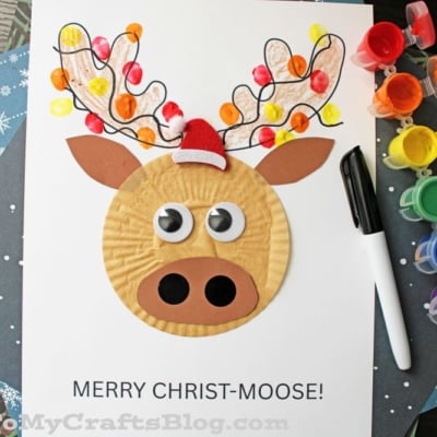 A cupcake liner moose head design, in the center of white cardstock paper, covered with Christmas lights made from a child's thumb dipped in craft paint