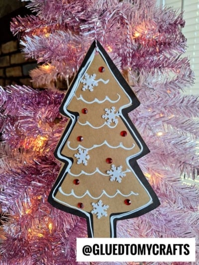 A handmade Paper Gingerbread Tree Craft ornament with white designs, red gems, and snowflake shapes hangs on a pink tinsel tree. The handle @GLUEDTOMYCRAFTS is visible at the bottom.