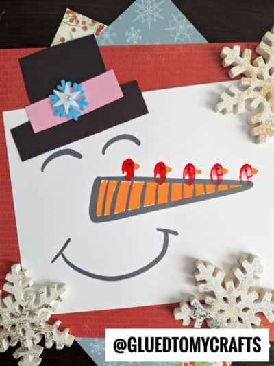 A snowman face drawing with a black top hat, orange carrot nose, smiling mouth, and four fingerprint birds perched on the nose, surrounded by paper snowflakes and craft paper. @GLUEDTOMYCRAFTS is at the bottom.