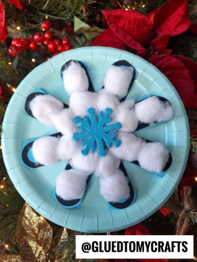 A blue paper plate features a cotton ball snowflake craft made from black-painted spoons, fluffy cotton balls, and a blue foam snowflake, displayed before red poinsettias and holiday greenery. @GLUEDTOMYCRAFTS is at the bottom.