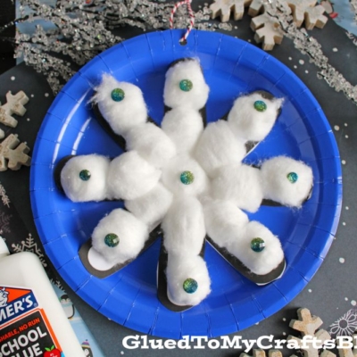 A blue paper plate displays a cotton ball snowflake ornament, decorated with blue glittery dots. A bottle of glue and assorted craft supplies are nearby.