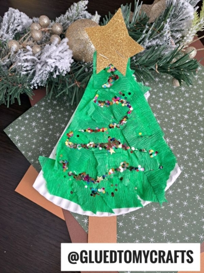 A handmade Torn Paper Christmas Tree craft with green paper, glitter, and a gold star topper, placed on holiday cardstock, with festive greenery in the background. The text @GLUEDTOMYCRAFTS is at the bottom.