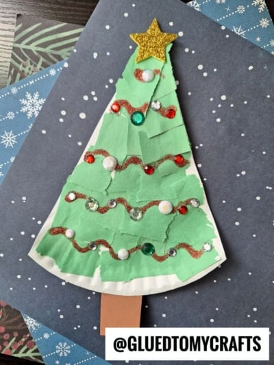 A handmade Torn Paper Christmas Tree craft made from green paper strips, decorated with sequins, white pom-poms, and a gold glitter star on top, plus a brown trunk, set against a dark background with white dots.