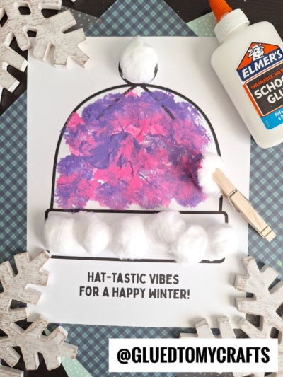 A pom pom stamped winter hat coloring page decorated with purple and pink paint, topped with cotton balls. The page reads “HAT-TASTIC VIBES FOR A HAPPY WINTER!” Glue, clothespin, and paper snowflakes surround it.