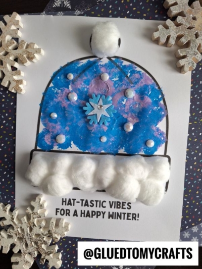 A child’s blue and purple painted winter hat craft, featuring cotton balls and pom-poms, is inspired by a fun pom pom stamped winter hat technique and placed on a decorated background with white snowflake ornaments. Text reads HAT-TASTIC VIBES FOR A HAPPY WINTER!.