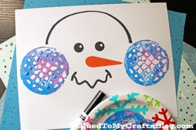 A snowman face printable is decorated with blue and purple potato masher stamped ear muffs using paint. Craft supplies, snowflake ornaments, and a black marker are nearby