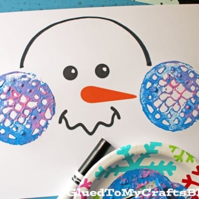 A snowman face printable is decorated with blue and purple potato masher stamped ear muffs using paint. Craft supplies, snowflake ornaments, and a black marker are nearby