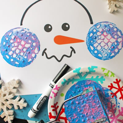 A snowman face printable is decorated with blue and purple potato masher stamped ear muffs using paint. Craft supplies, snowflake ornaments, and a black marker are nearby. Text reads, “FREE PRINTABLE!!!”.