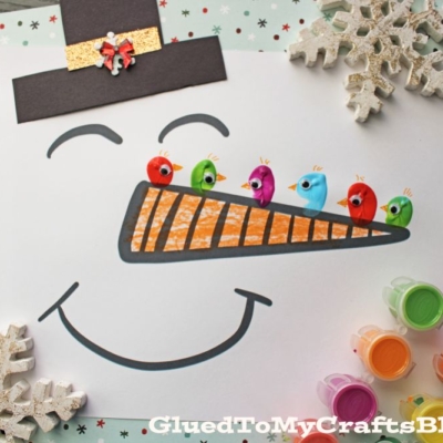 A snowman face drawing with a carrot nose, decorated with fingerprint birds and colorful pom-poms. Snowflakes and paint pots surround the paper.