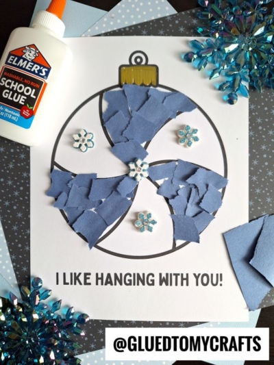 A torn paper christmas ornament craft featuring blue paper scraps glued onto a printed ornament outline, decorated with sparkling snowflake embellishments. Elmer’s glue, blue paper scraps, and sparkling snowflake decorations are nearby.
