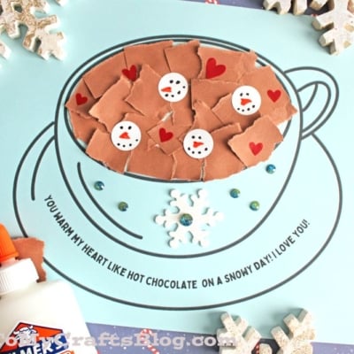 A blue paper displays a torn paper hot chocolate mug, filled with brown pieces and snowman faces, decorated with red hearts and white snowflakes. Elmers glue and snowflake ornaments sit nearby.