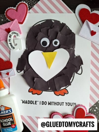 A handmade torn paper penguin Valentine craft features a white heart belly, yellow beak, googly eyes, and orange feet. Surrounded by felt hearts, envelopes, a glue bottle, and a striped paper background.