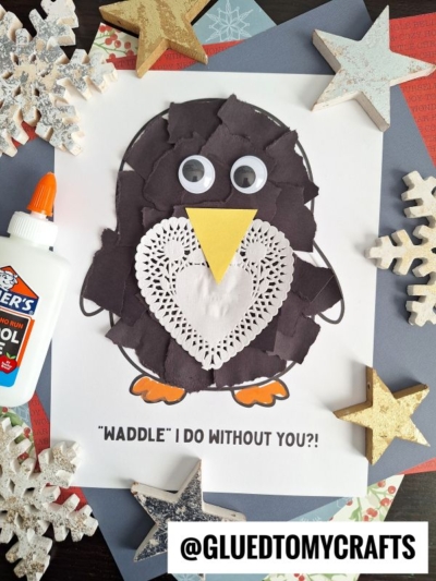 A torn paper penguin craft made from torn black paper, a paper doily, googly eyes, and a yellow beak sits on a decorated page with WADDLE I DO WITHOUT YOU?! text and is surrounded by snowflakes and stars.