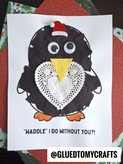 A torn paper penguin craft made of black paper, a yellow triangle beak, a white heart doily belly, googly eyes, and a red Santa hat. Text below reads, “WADDLE I DO WITHOUT YOU?!” @GLUEDTOMYCRAFTS.