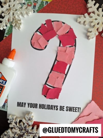 A holiday craft featuring a torn paper candy cane outline filled with pink and red paper pieces, with the text “May your holidays be sweet!” A bottle of glue and decorative snowflakes are nearby.