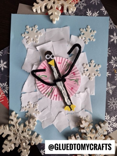 A Popsicle Stick Snow Tubing Penguin made from a clothespin, pipe cleaner, googly eyes, and paper stands on a snowy craft scene with torn white paper, snowflakes, and a pink cupcake liner on a blue background.