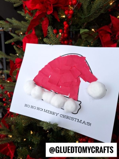 A handmade torn paper Santa hat craft made with red paper pieces and cotton balls is displayed on white paper. The craft sits in front of a decorated Christmas tree with red ribbons and ornaments. Text reads @GLUEDTOMYCRAFTS.