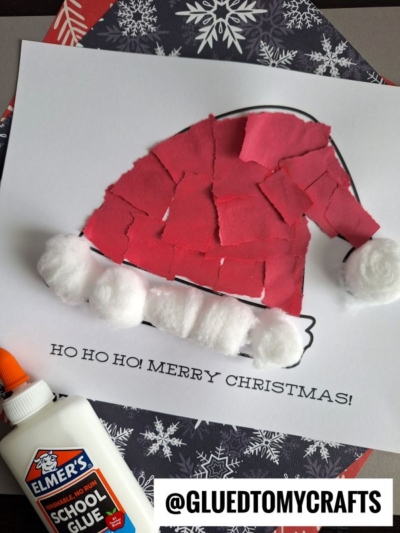 A torn paper Santa hat craft made with red paper pieces and cotton balls, glued to an outlined hat on paper that says HO HO HO! MERRY CHRISTMAS! Elmers glue and patterned paper are nearby.