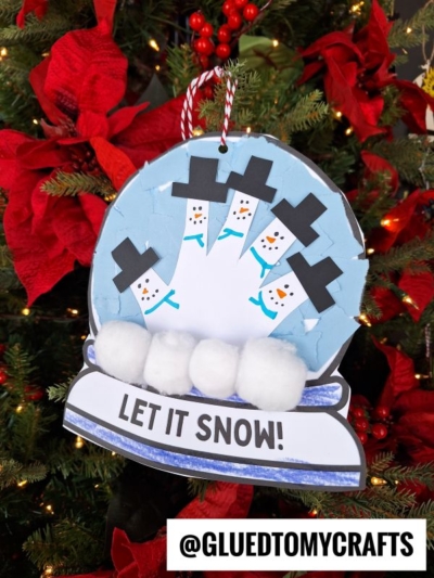 A handmade torn paper snow globe ornament featuring five snowmen made from a traced handprint, with cotton balls as snow, hangs on a Christmas tree decorated with red poinsettias. Text reads LET IT SNOW! and @GLUEDTOMYCRAFTS.