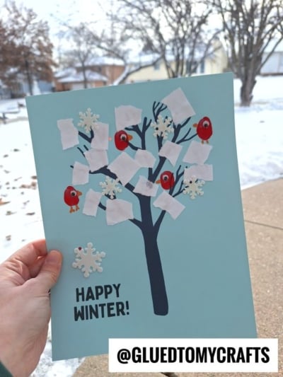 A hand holds a blue paper featuring a torn paper winter tree adorned with cutout snowflakes, red birds, and white tissue snow. HAPPY WINTER! is written below. A snowy neighborhood is visible in the background.